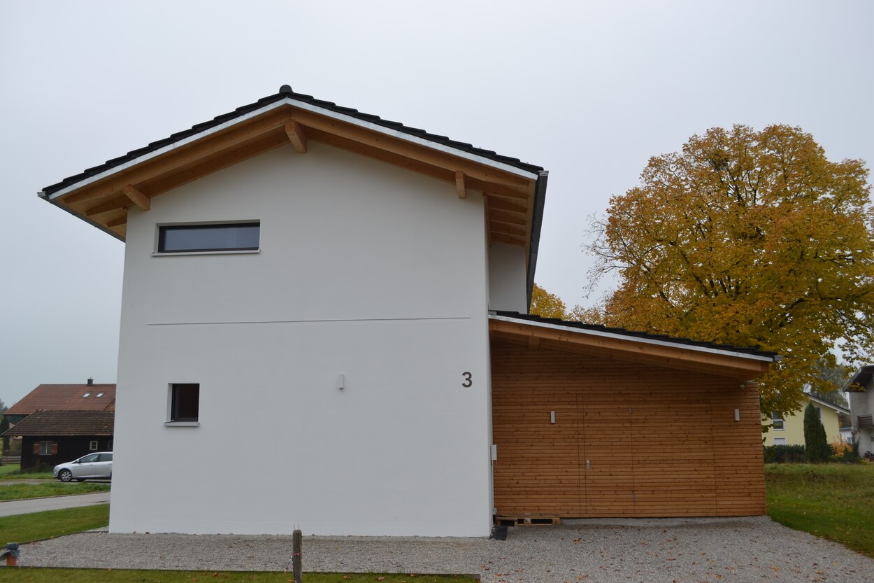 Ein modernes Passivhaus mit weißer Fassade und Holzverkleidung steht neben einem herbstlich gefärbten Baum in Übersee.