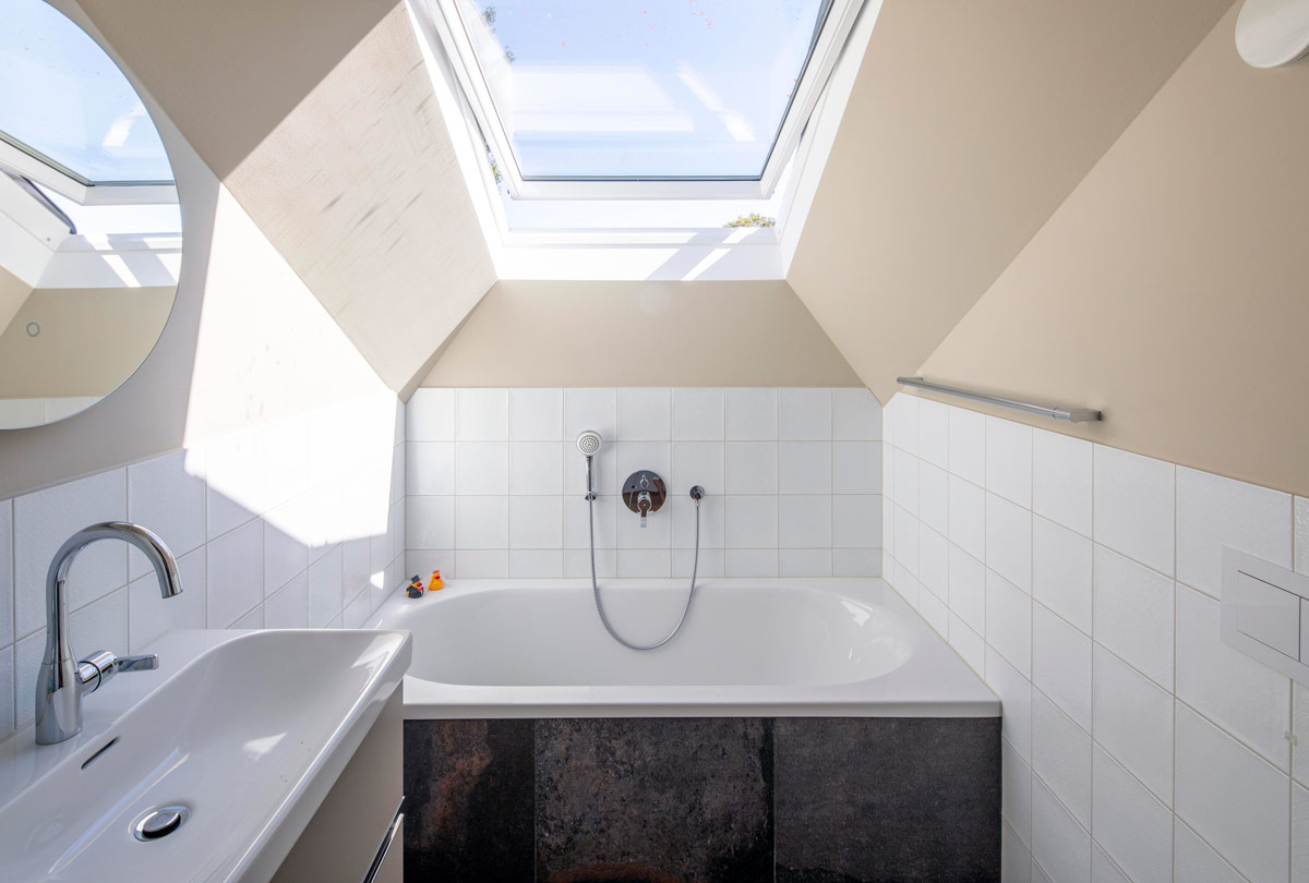 Ein modernes Bad im Dachgeschoss mit Badewanne unter einem großen Dachfenster, das viel Tageslicht hereinlässt.