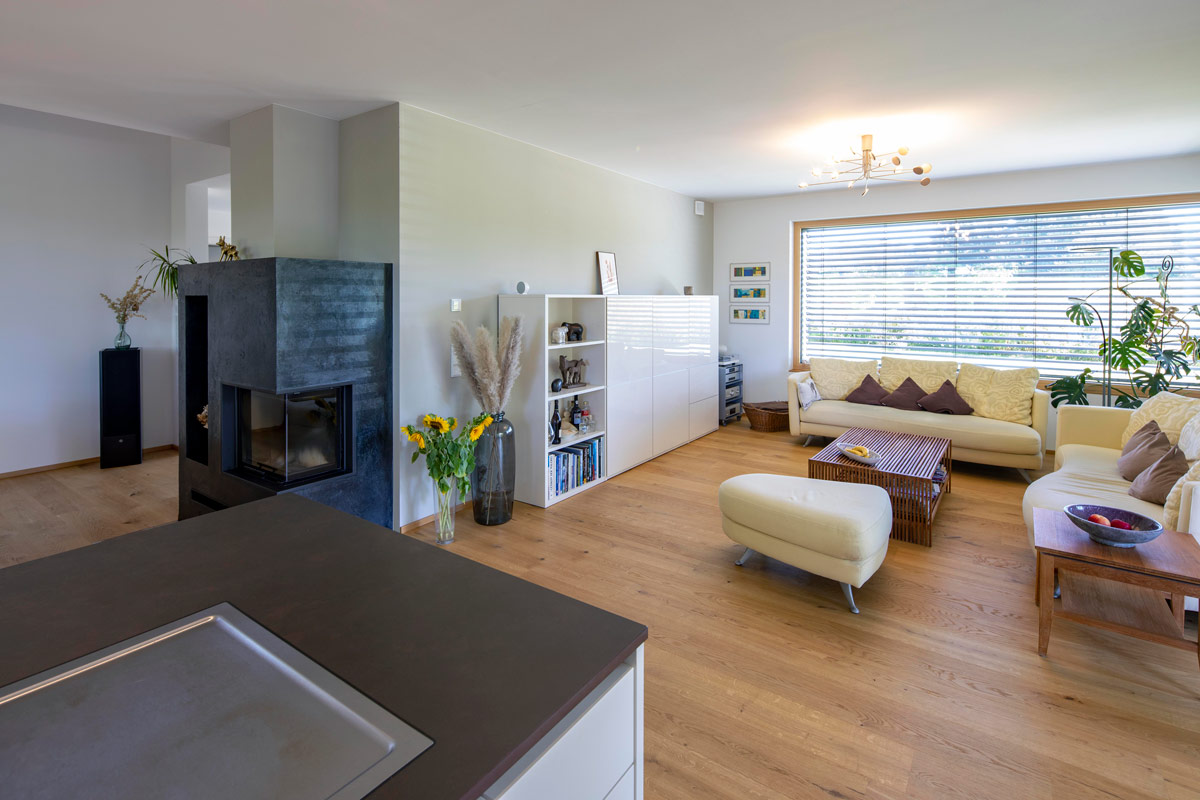 Ein modernes Wohnzimmer in einem Holzhaus in Glonn mit stilvollen Möbeln, Kamin und großen, lichtdurchfluteten Fenstern.