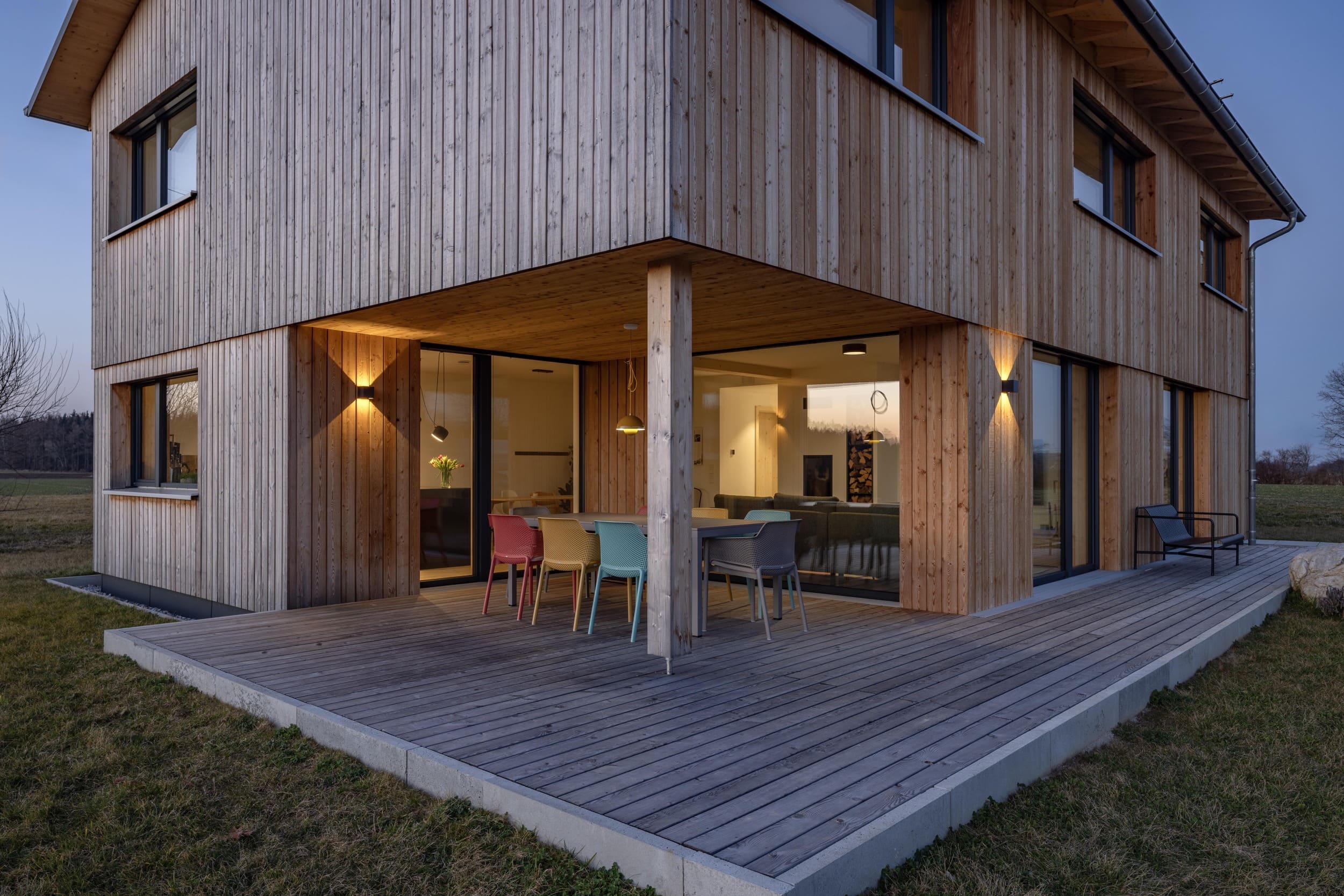 Ein modernes Holzhaus mit bunten Stühlen steht bei Abenddämmerung in einer ruhigen, ländlichen Umgebung.