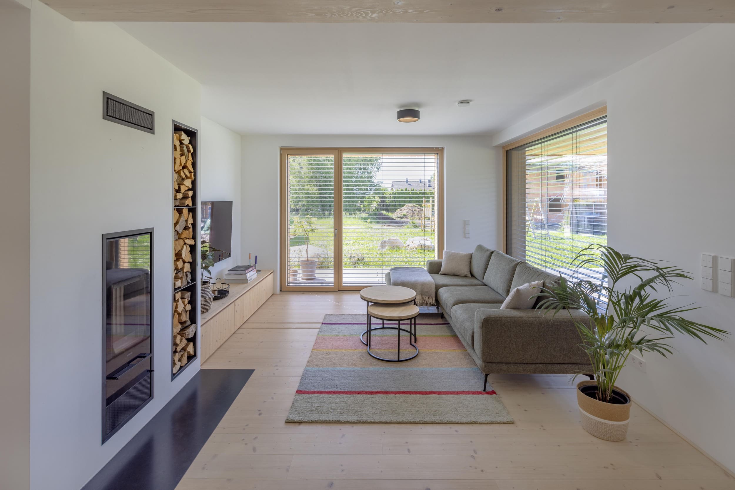 Ein helles Wohnzimmer mit grauem Sofa, Couchtischen und großen Fenstern bietet einen weiten Blick auf den Garten.