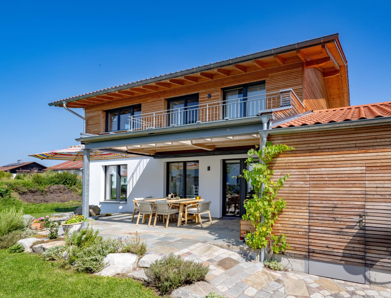 Ein modernes Holzhaus mit Terrasse und Gartenmöbeln steht unter einem strahlend blauen Himmel.
