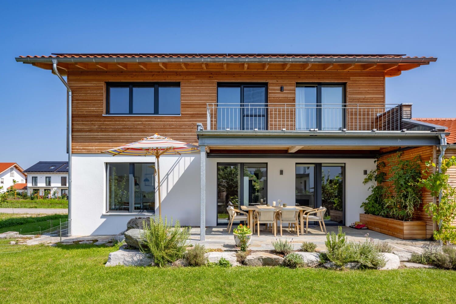 Ein modernes zweistöckiges Haus mit Holzelementen und einem einladenden Garten bietet eine gemütliche Terrasse zum Verweilen.