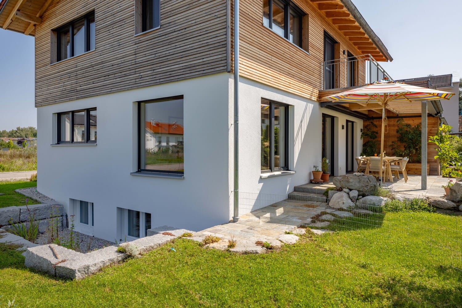 Ein modernes Haus mit Holzfassade und Terrasse im Garten lädt bei Sonnenschein zum Entspannen unter dem Sonnenschirm ein.