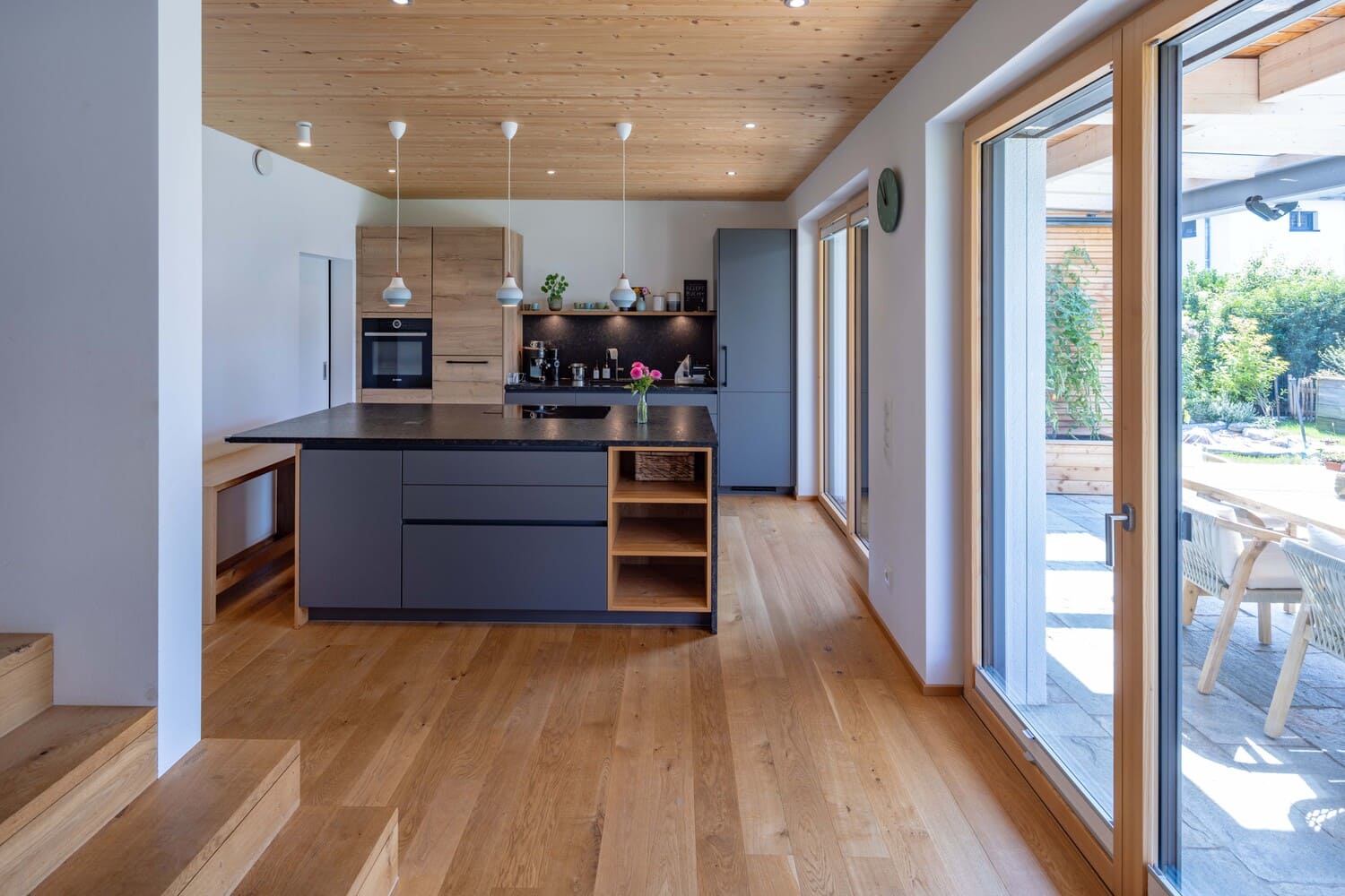 Moderne Küche mit Holzdecke, grauer Insel und großen Fensterfronten, die viel Tageslicht in den Raum lassen.