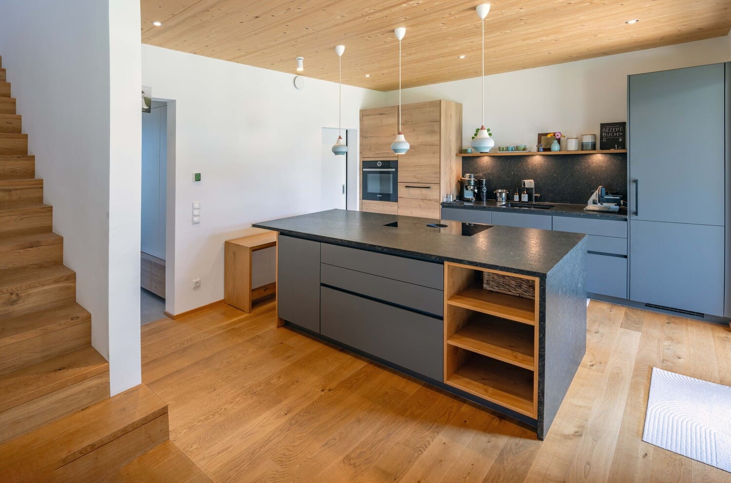 Eine moderne Küche mit Holz-Elementen und grauer Insel neben der Treppe verleiht dem Raum ein elegantes Ambiente.