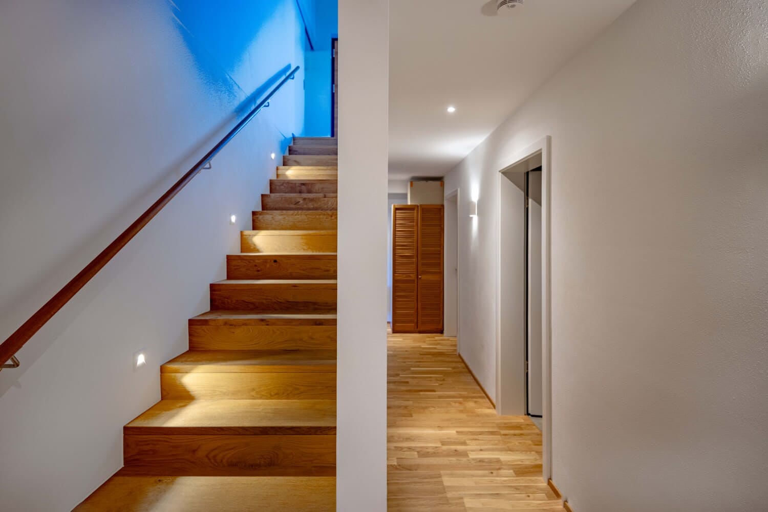 Eine beleuchtete Holztreppe führt in einen hellen, minimalistischen Flur eines modernen Interieurs.
